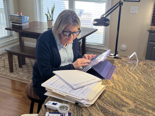 Rosanna in her home. She sits at a round wooden table, working on a tablet. She looks at a bold-lined notepad with large print.
