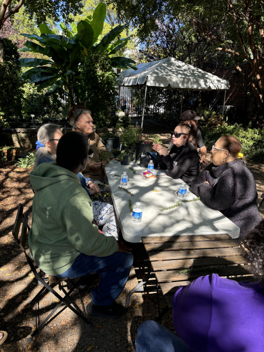 A group of people sit around an outdoor table as a man discusses various herbs and passes samples for the group to explore.