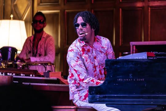 Matthew Whitaker plays a black grand piano. He wears a red and purple flower-patterned shirt. Matthew has short dreadlocks and a goatee. In the left background the drummer is visible.